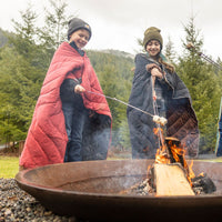 Two children wrapped in puffy camping blankets roasting marshmallows over a fire pit outdoors