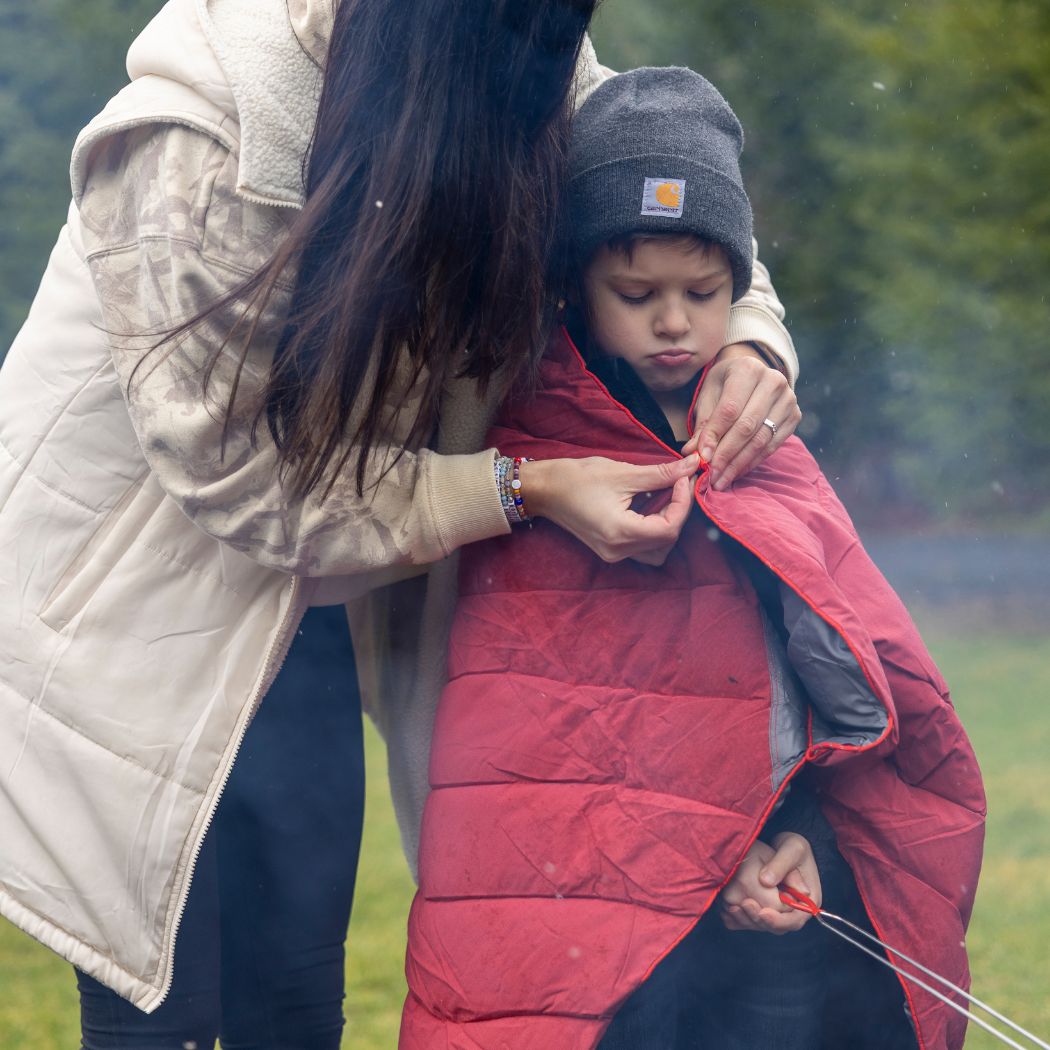 Child wrapped in red puffy blanket outdoors near campfire, adult helping zip it closed