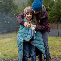 Child wrapped in teal puffy camping blanket being helped by adult outdoors in misty forest