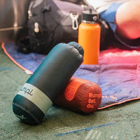 Rolled travel blanket in dark teal beside a red variant, water bottle, and backpack on carpet