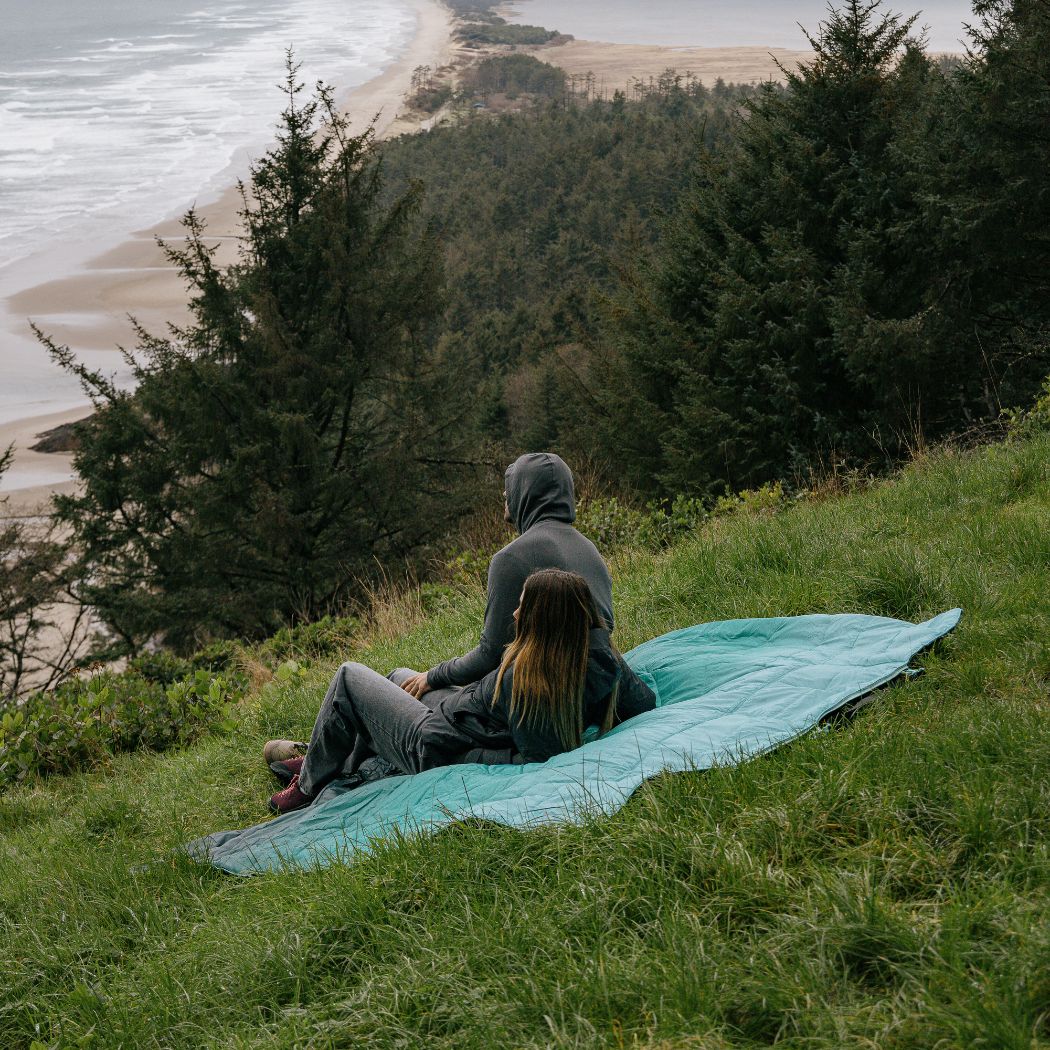 Teal gradient outdoor mat with two people sitting on grassy hillside overlooking coastline
