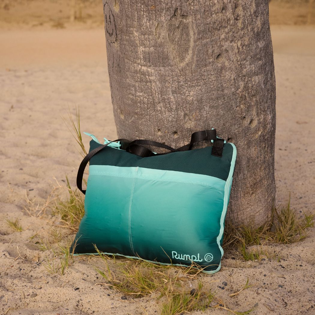 Teal gradient outdoor mat with black straps leaning against palm tree trunk on sandy beach