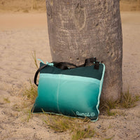 Teal gradient outdoor mat with black straps leaning against palm tree trunk on sandy beach
