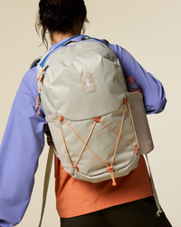 Light colored daypack with orange cord details worn by person in purple jacket