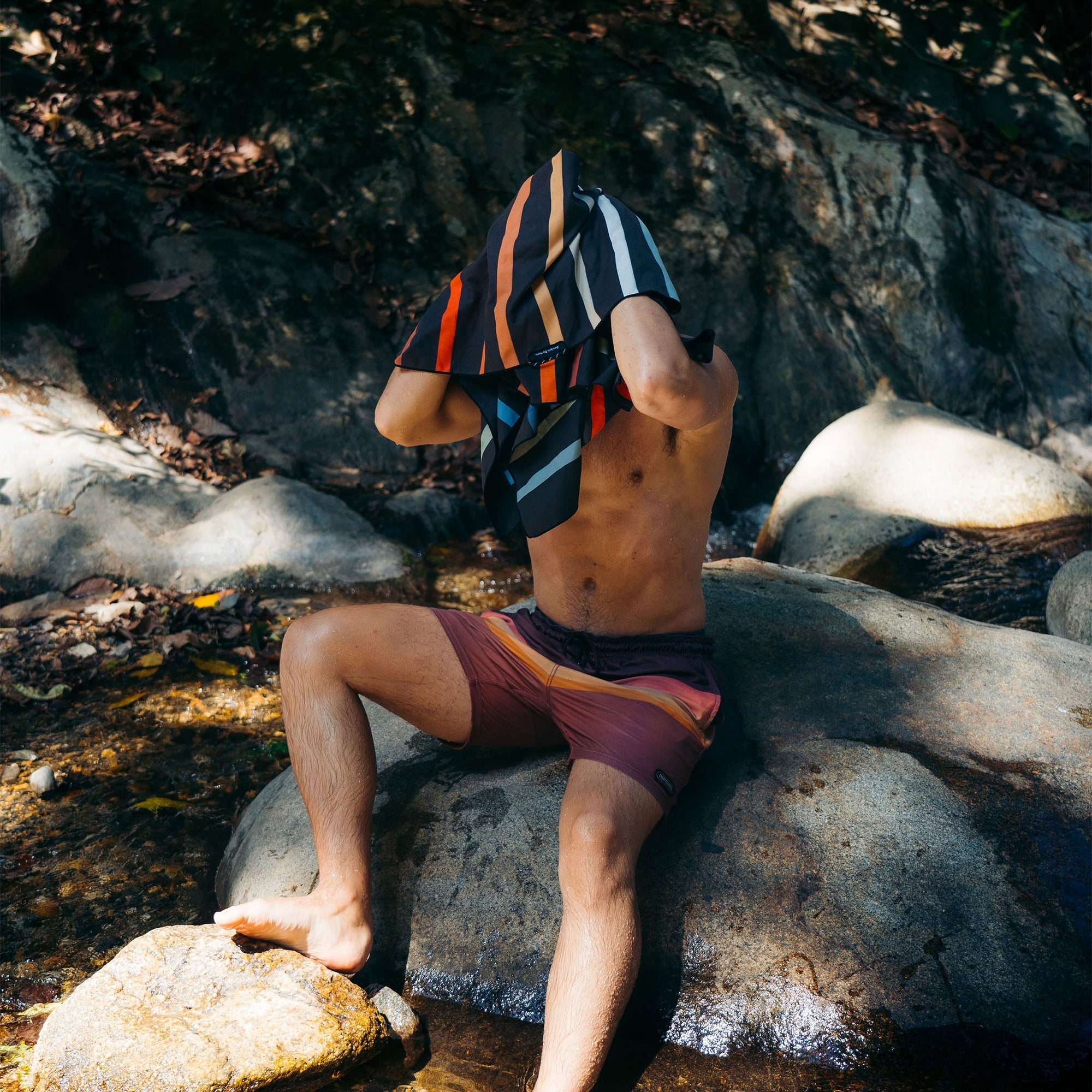 Striped quick-dry towel being held up by person sitting on rocky riverbank